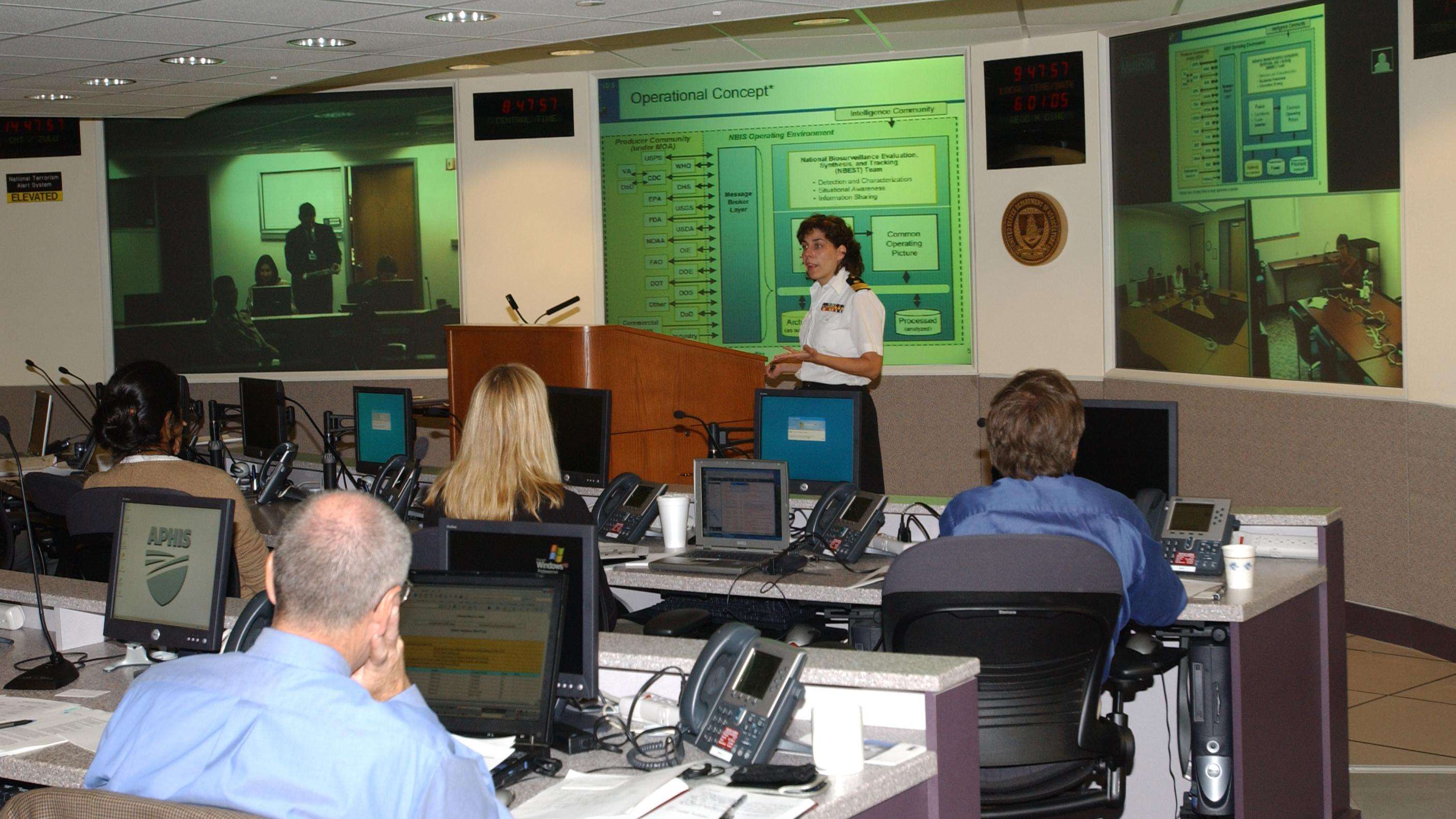 A woman in uniform stands between a podium and projected image titled "operational concept". People sitting in rows of desks with computers and notebooks watch the woman.