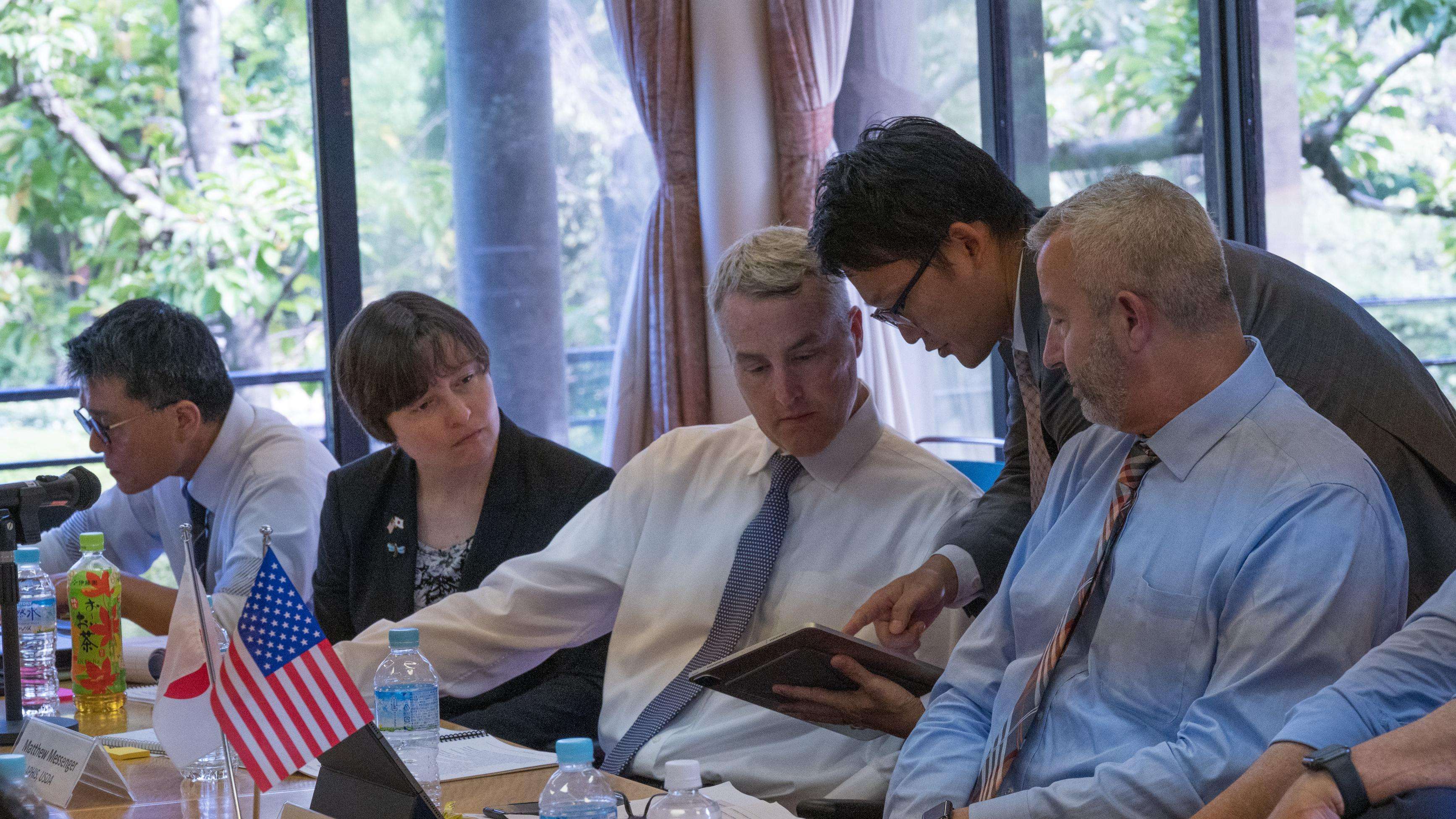 Group of people sitting at a table in conversation with notebooks and a U.S. flag on table.