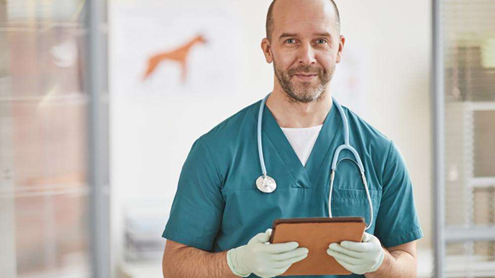 A man with green scrubs, a clipboard, and medical gloves holds a clipboard and looks at the camera.