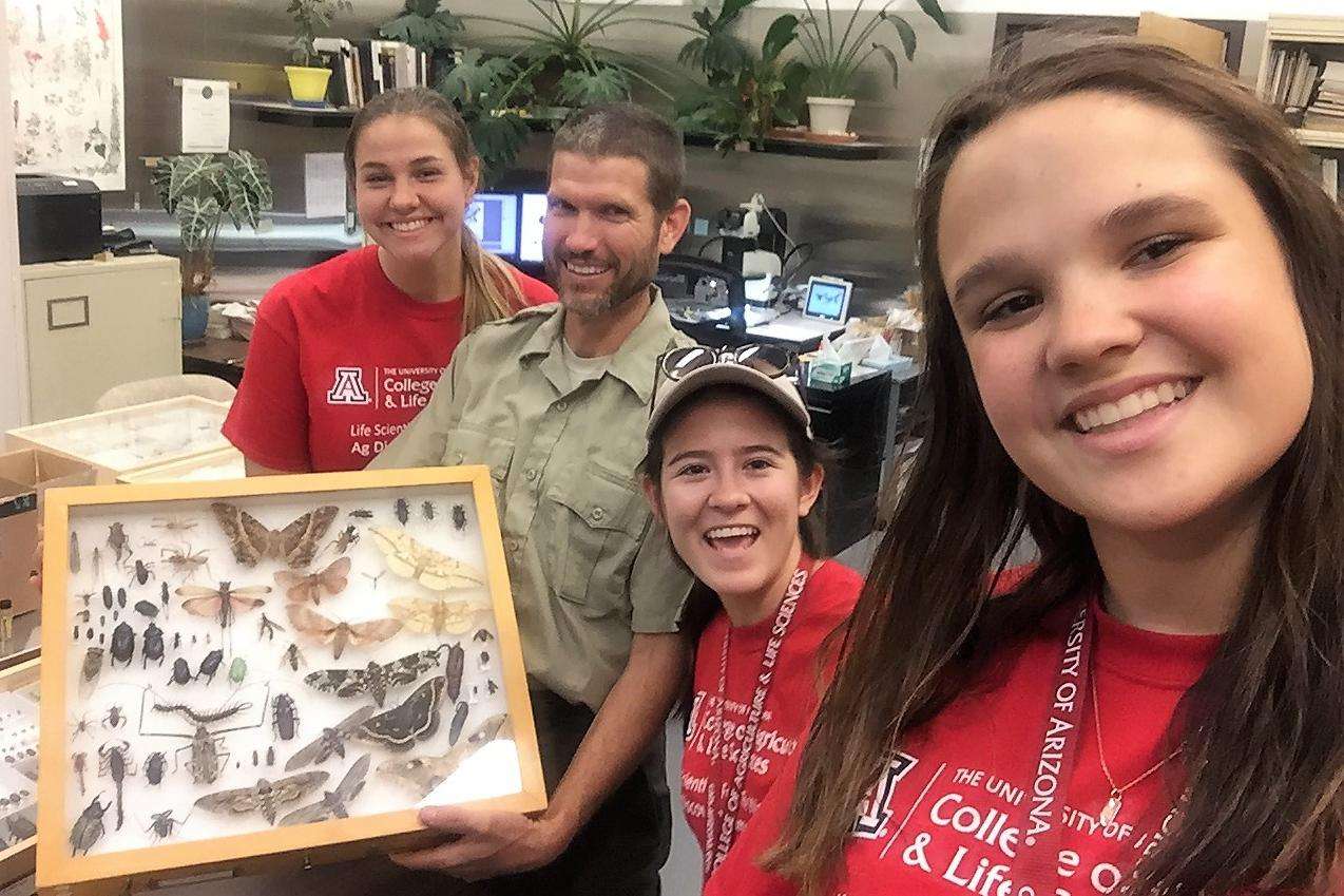 Three female students pose for a photo with an APHIS entomologist and his insect collection.