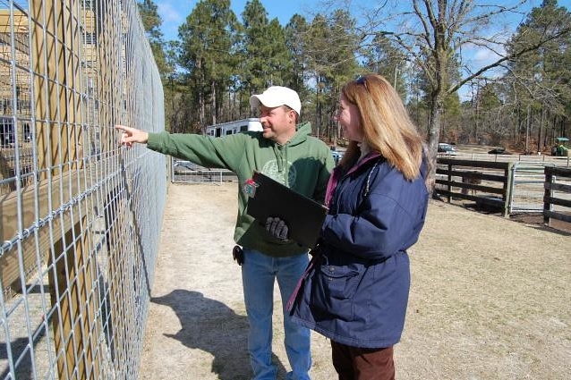 Female veterinarian holding a clipboard. She's standing next to a man pointing towards a fenced enclosure.