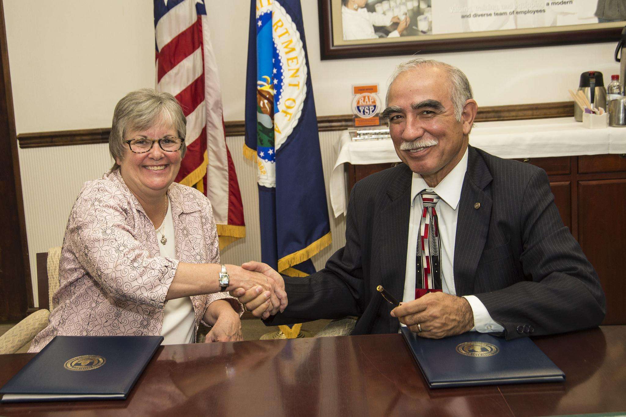 Woman and man sitting a table in front of USDA and U.S. flags shaking hands