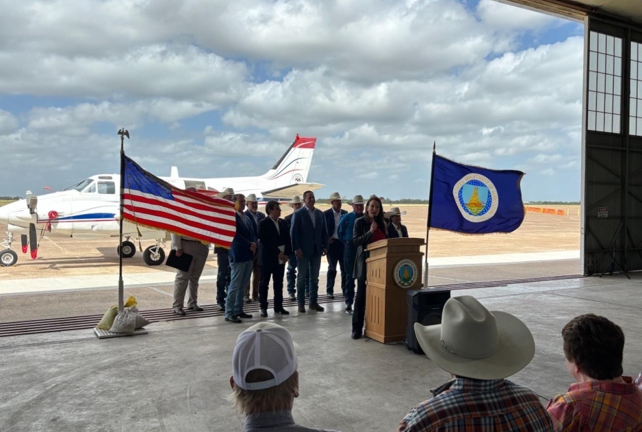 Secretary Rollins at the podium with a group of people behind and a plane in the background